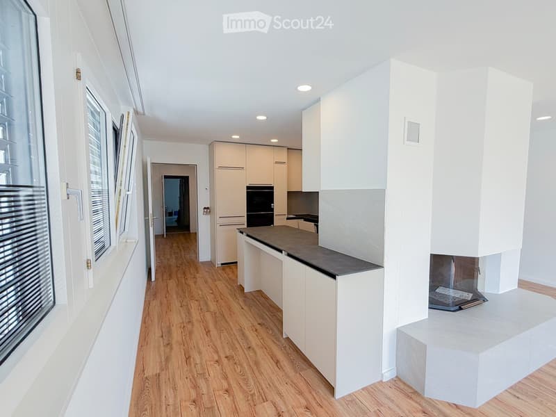Modern, minimalist kitchen with white cabinets, a dark countertop, and a built-in oven. The space features light wooden flooring and large windows with blinds.