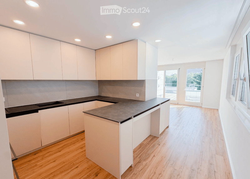 Modern kitchen with light-colored cabinets, a black countertop, and a wooden floor. Large windows and a sliding glass door lead to a balcony.