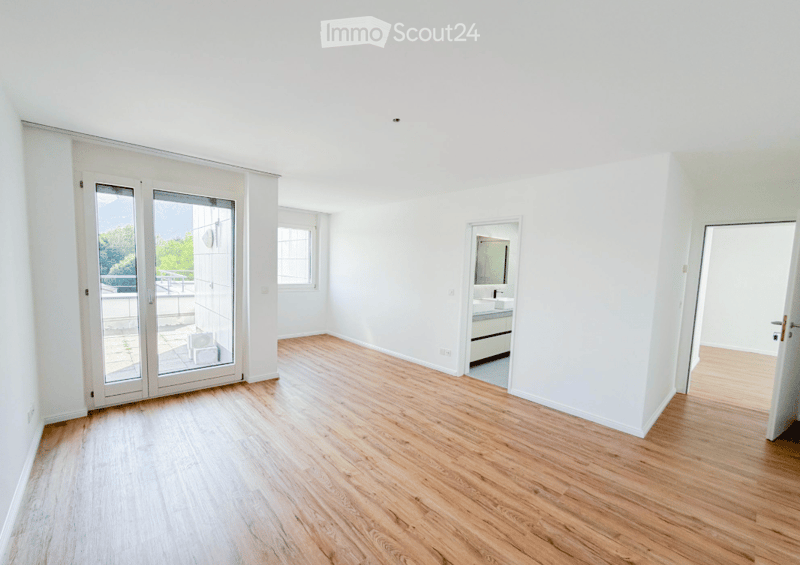 Bright, unfurnished room with wooden flooring, large glass sliding doors leading to a balcony, and a small adjacent bathroom.