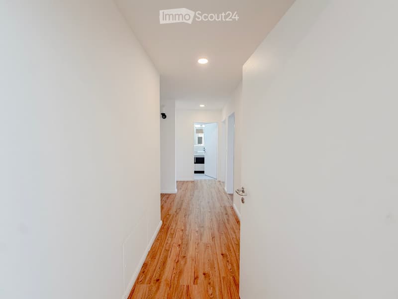 Empty hallway with white walls, wooden flooring, and recessed lighting. A door at the end leads to another room.