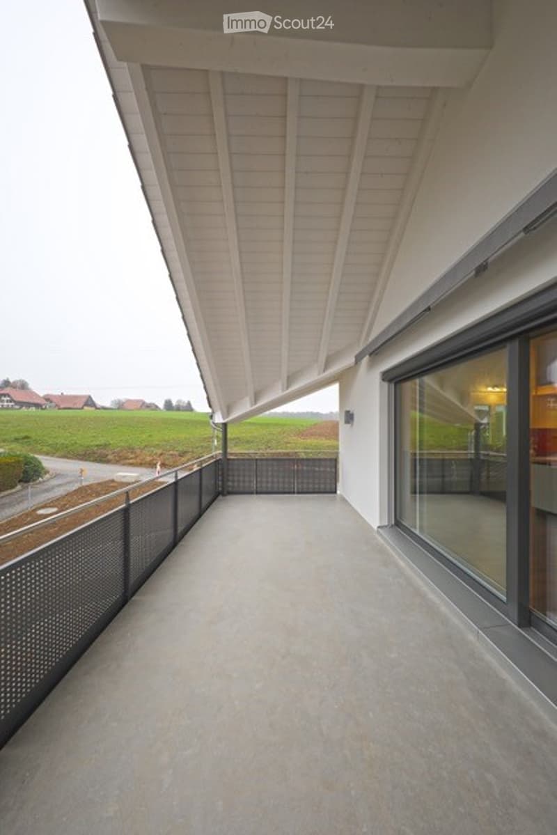Modern balcony with a sleek white ceiling, large glass windows, and a view of green fields and a road.