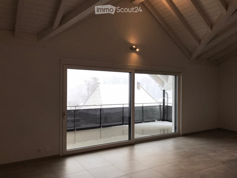 Empty attic room with exposed wooden beams, large sliding glass doors leading to a balcony, tiled floor, and modern lighting fixture.