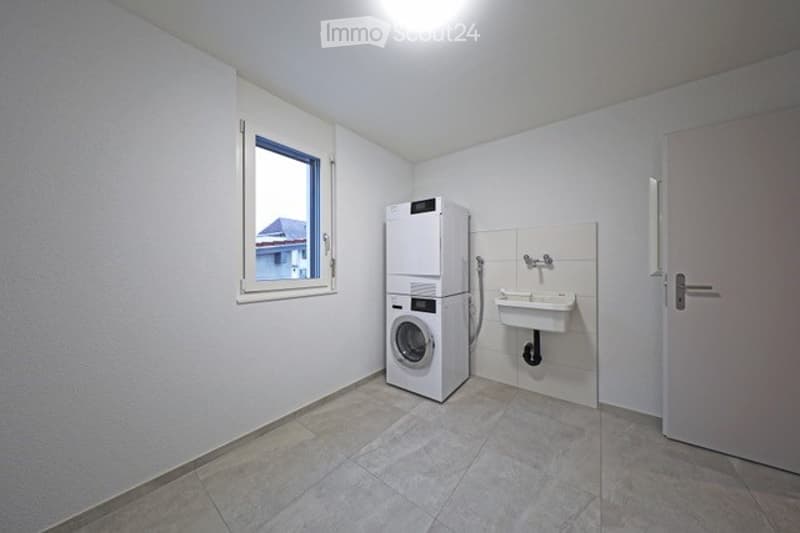 A small, clean bathroom with a washing machine, a sink, and a window offering a view of the outside.