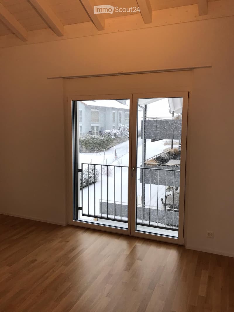 Empty room with wooden flooring and large sliding glass doors leading to a balcony with a metal railing. The view shows a snowy outdoor area with a neighboring building and some greenery.