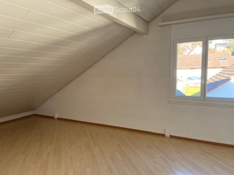 Empty attic room with wooden flooring, white walls, and a large window offering a view of the outside. The ceiling has exposed wooden beams.