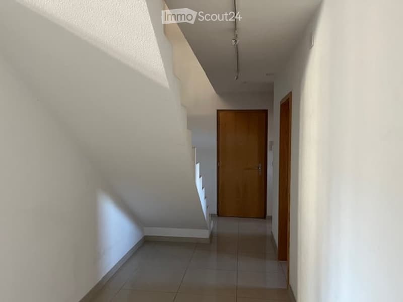 A clean, modern hallway with white walls, tiled flooring, and a wooden door at the end. The hallway features a staircase leading upwards, with a simple, contemporary design.
