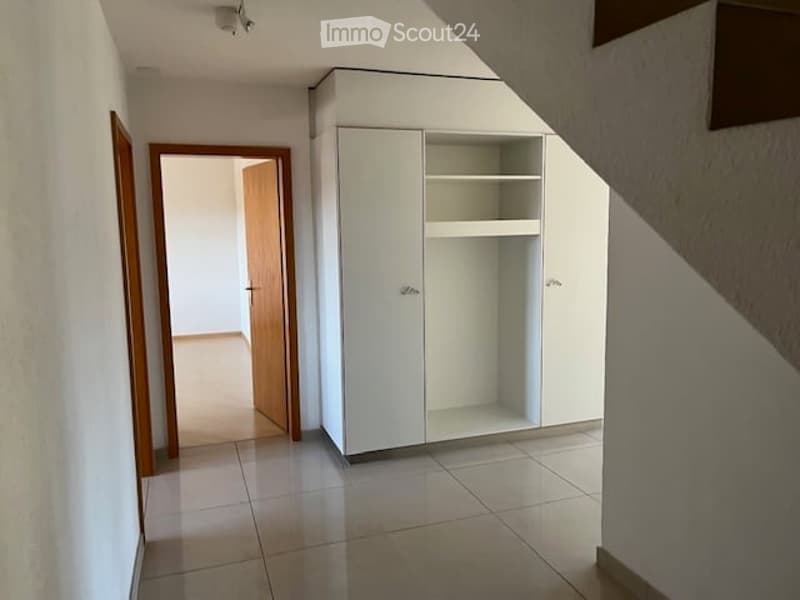 Empty hallway with tiled floor, white walls, and a white built-in closet with empty shelves. A wooden door leads to another room.