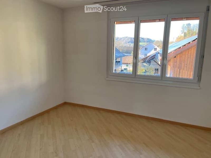 Empty room with wooden flooring and large window showing outdoor view.