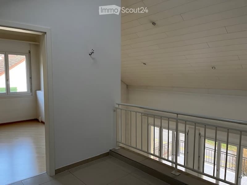 Empty hallway with wooden flooring, white walls, and a metal railing leading to another room. The area is well-lit with natural light coming from a window on the left side.