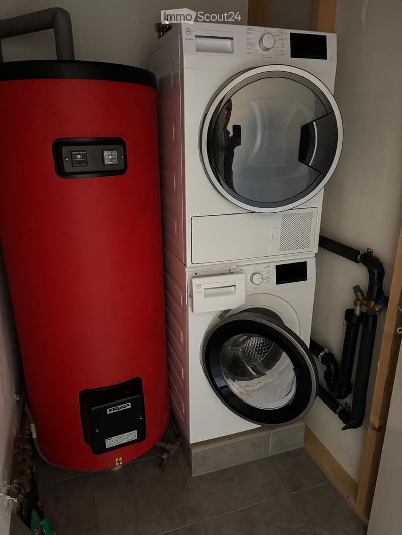 A compact utility room with a white washing machine, a matching dryer, and a red water heater. The room has tiled flooring and visible plumbing.
