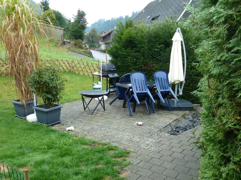 Outdoor patio with a table, chairs, umbrella, grill, and potted plants, surrounded by greenery and a wooden fence.