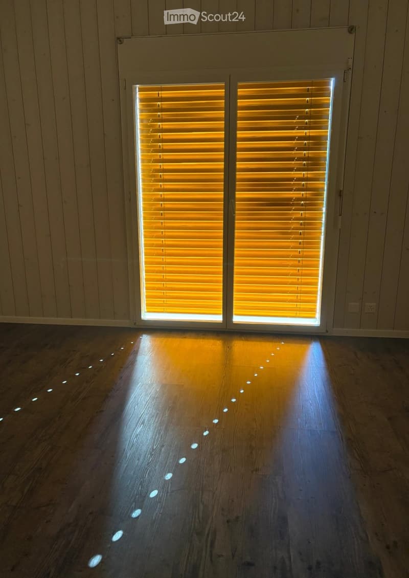 Empty room with wooden flooring and a large sliding glass door with blinds. The floor has small, evenly spaced LED lights embedded in it.
