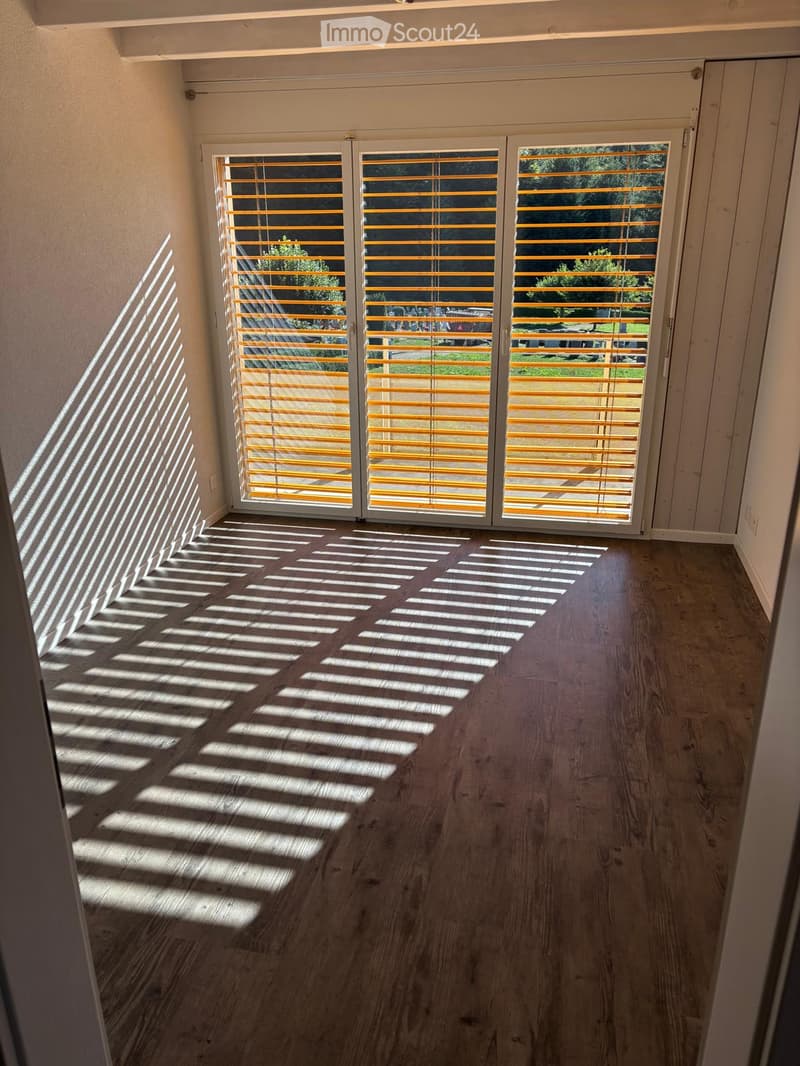 Empty room with wooden flooring and large sliding glass doors with yellow blinds, allowing natural light to create striped shadows on the floor.