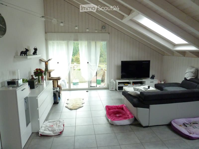 Bright living room with white walls, slanted wooden ceiling, large window with white curtains, sofa, coffee table, cat tree, cat beds, television, and decorative items.