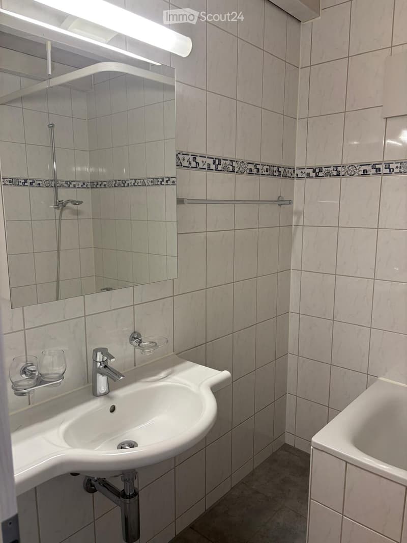 Modern bathroom with a white sink, chrome faucet, mirror, and bathtub. The walls are tiled with a decorative border, and there are two cups on a shelf next to the sink.