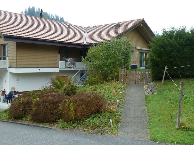 Two-story house with a tiled roof, wooden siding, and a balcony. The house is surrounded by greenery, including bushes, trees, and a small garden area with a wooden fence and a pathway leading to the entrance.