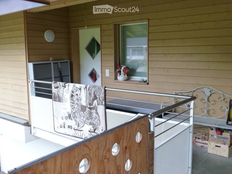 Balcony with wooden railing, bench, towel, vase on window sill, and a small storage area with boxes.