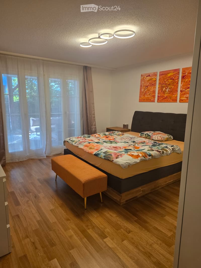 Modern bedroom with wooden flooring, large bed with floral bedding, matching stool, nightstand, and abstract paintings on the wall.