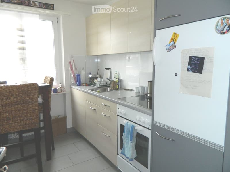 Modern kitchen with light-colored cabinets, a refrigerator, an oven, a sink, and a dining area with a chair and table.