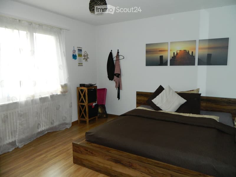 Cozy bedroom with wooden bed, nightstand, and large window with white curtains. Features decorative wall art and a coat rack with hanging items.
