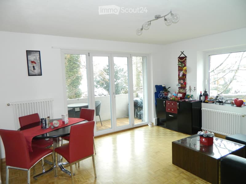 Bright living and dining area with wooden flooring, large windows, and a balcony access. Features a dining table with red chairs, a black cabinet, a sofa, and decorative items on the windowsill.