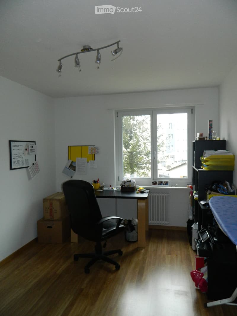 Office space with wooden floor, large window, desk, chair, shelf, radiator, and board on wall.