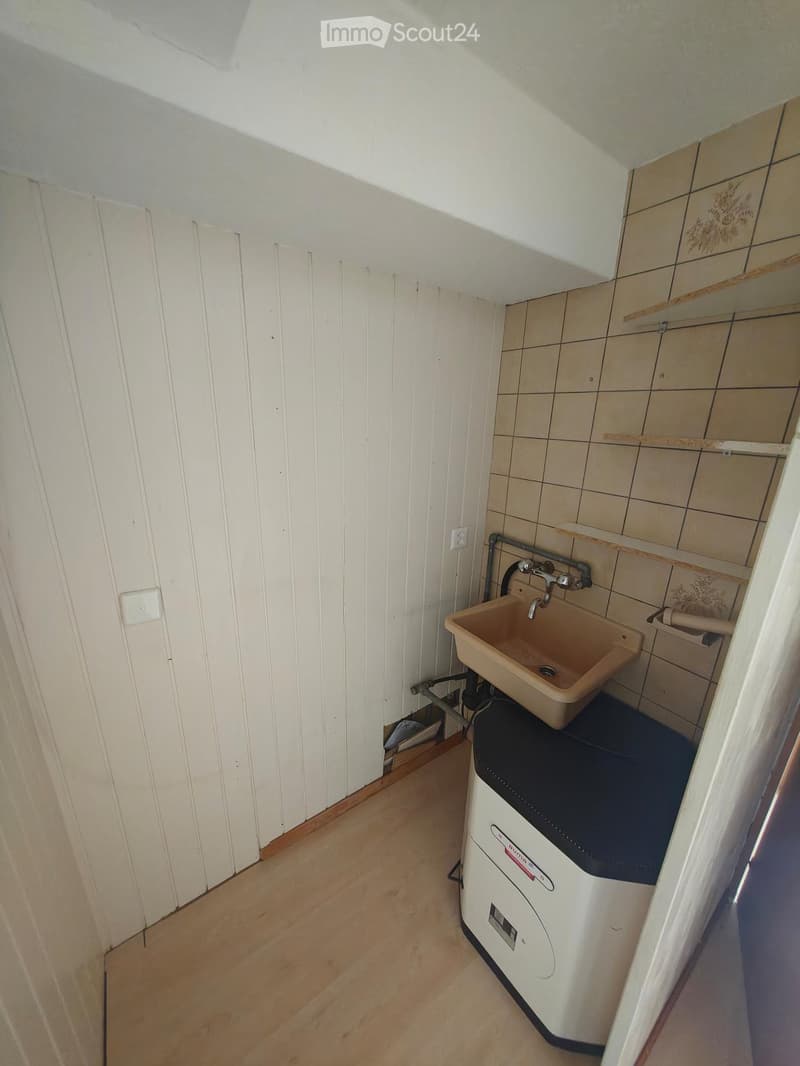Small bathroom with a sink, faucet, toilet, and washing machine. The walls are tiled, and the floor is wooden.