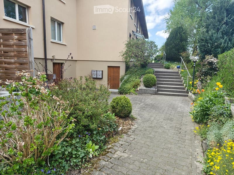 Garden path leads to a two-story house with beige walls, wooden doors, and windows. The garden is well-maintained with various plants, shrubs, and flowers, and includes a set of concrete steps leading up to the entrance.