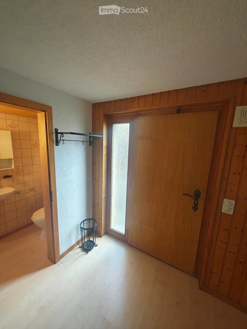 A narrow hallway with wooden walls and a light wooden floor, featuring a small black metal basket on the floor and a partially open door leading to a tiled bathroom with a toilet and sink visible.