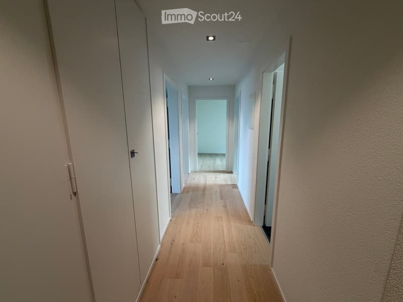 A long, narrow hallway with light wooden flooring, white walls, and recessed ceiling lights. Doors are visible on both sides, leading to other rooms.