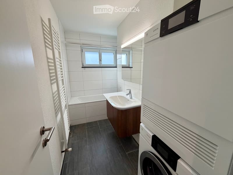 Modern bathroom with white tiles, a bathtub, a sink with a wooden cabinet, a washing machine, and a radiator.