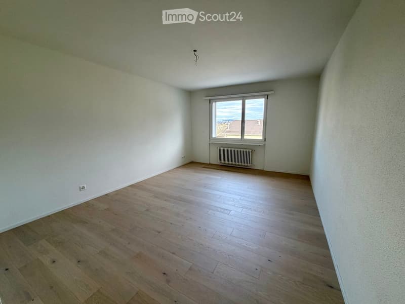 Empty room with wooden flooring, white walls, and a large window offering a view of the outside. The room includes a radiator below the window and a visible electrical outlet on the left wall.