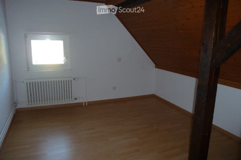 Empty attic room with wooden floor, white walls, and a single window letting in natural light. A radiator is mounted on the wall below the window.