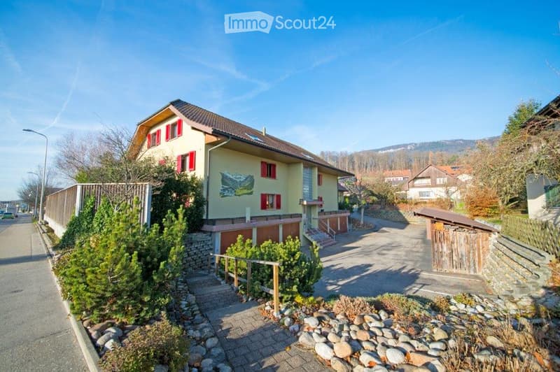 Two-story house with a yellow facade, red shutters, and a brown roof. The house features a small garden with shrubs and a stone pathway leading to the entrance.