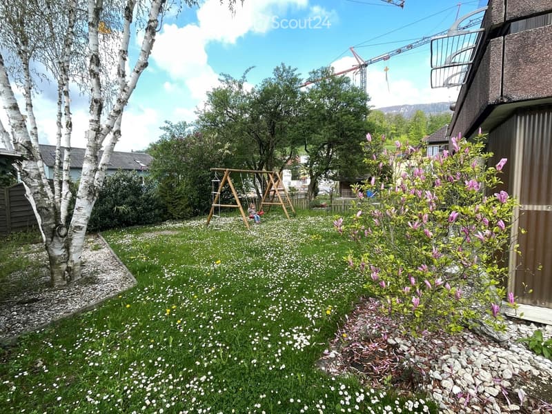 Lush garden with green grass, white flowers, a swing set, and surrounding trees and shrubs.