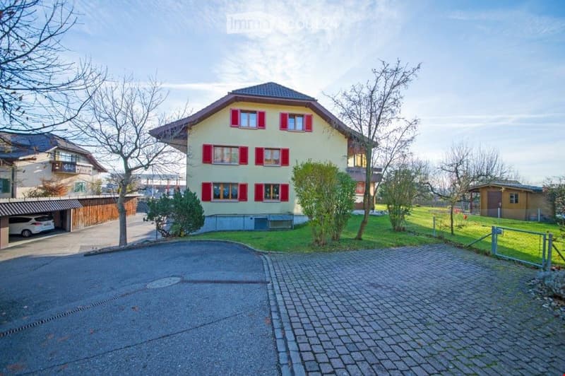 Two-story house with red shutters and a garage, surrounded by a well-maintained garden and driveway.