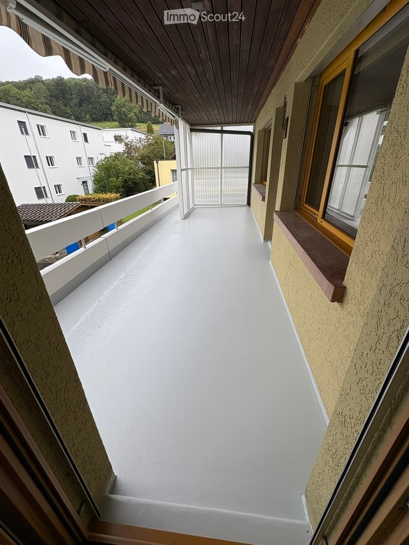 Balcony with white flooring, wooden ceiling, white railing, and a view of surrounding buildings and greenery.