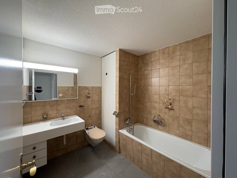 Modern bathroom with beige tiles, a white sink, a bathtub, a toilet, and a large mirror with built-in lighting.