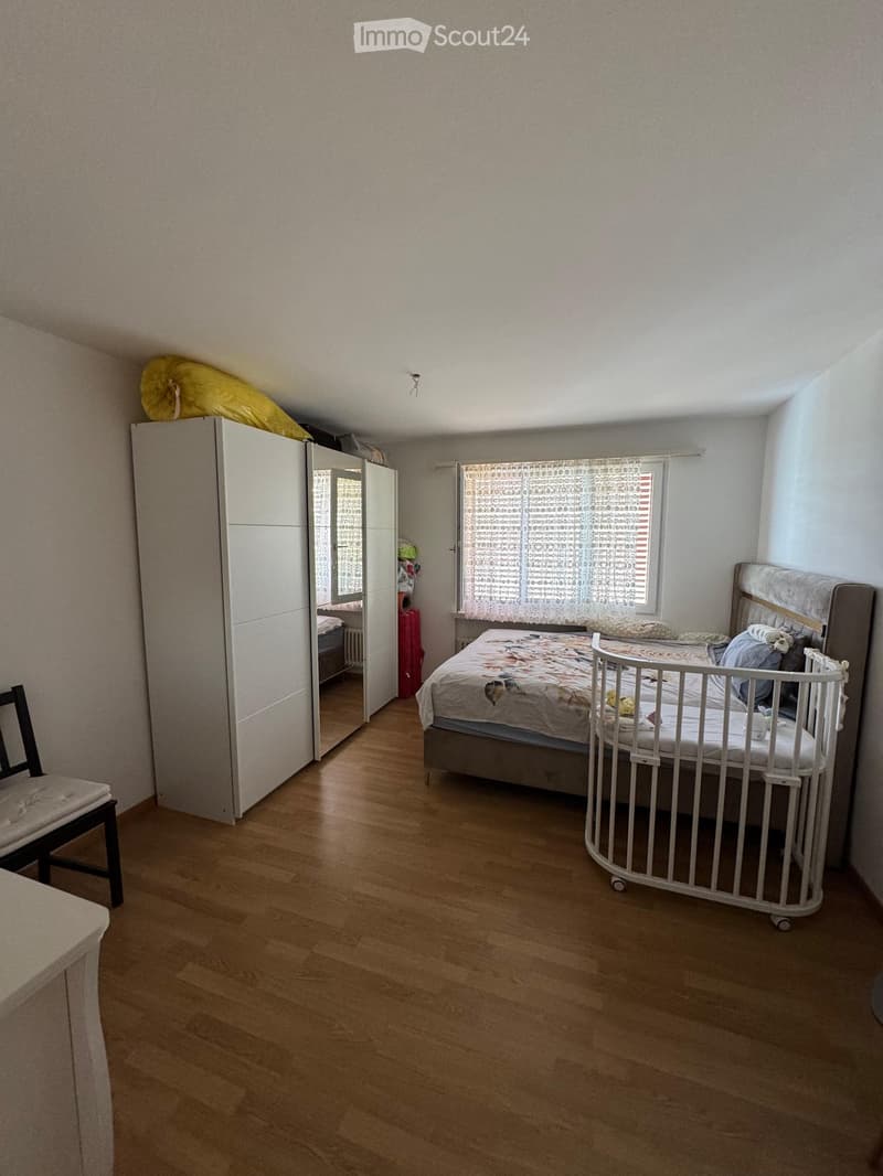 Bedroom with wooden flooring, a bed with a patterned duvet, a white wardrobe, a chair, and a baby cradle near the bed.