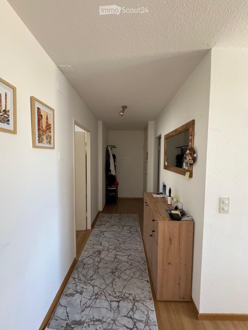 A narrow hallway with white walls, wooden flooring, and a patterned rug. Features include a wooden dresser with various items, a mirror, framed artwork, and a coat rack with hanging clothes.