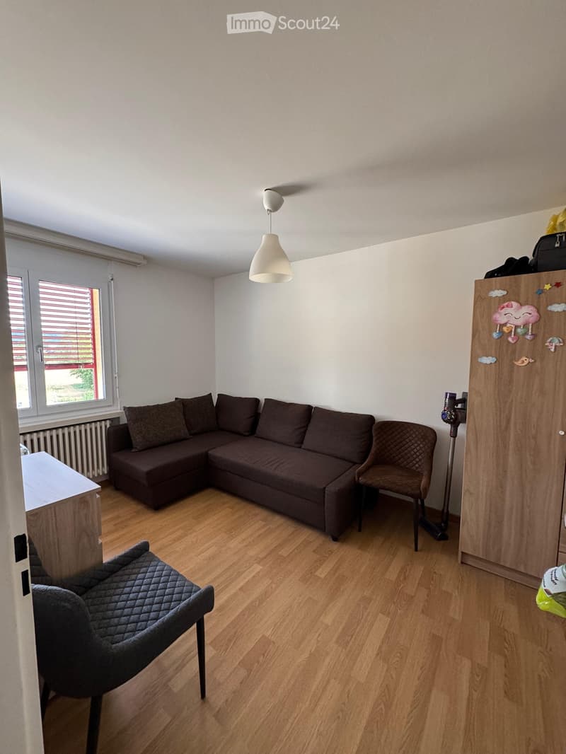 A cozy living room with a dark brown sectional sofa, a matching armchair, a small table, and a hanging lamp. The room features wooden flooring and large windows with blinds.
