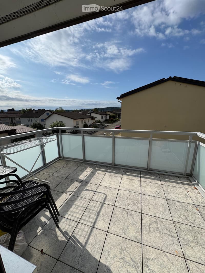 Balcony with tiled floor, glass railing, and a black wicker chair. The view shows a suburban neighborhood with houses and a clear sky.