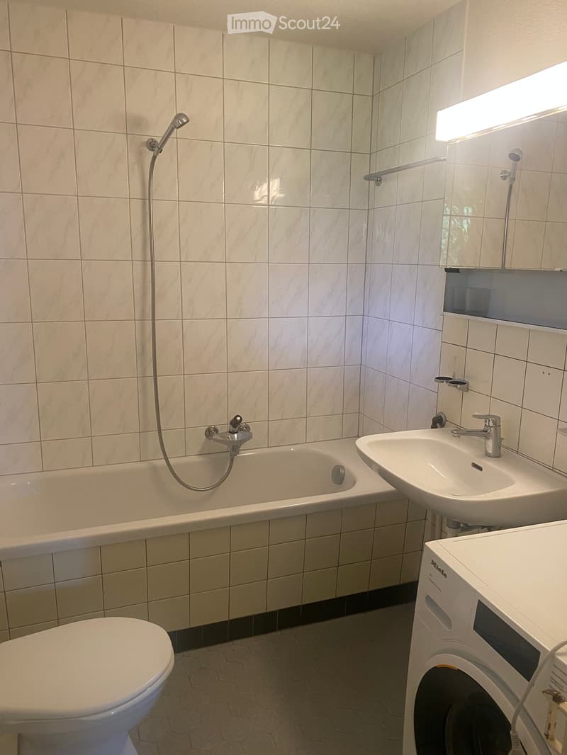Modern bathroom with white tiled walls, a bathtub with a handheld shower, a sink with a modern faucet, a toilet, and a washing machine in the corner.