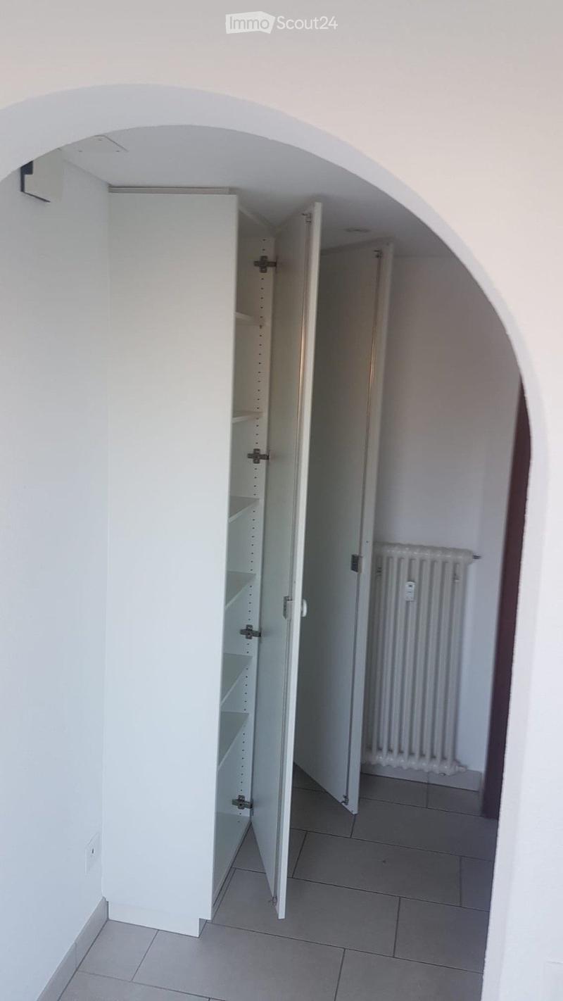 Empty hallway with white walls, tiled floor, open closet with empty shelves, and a radiator in the adjacent room.