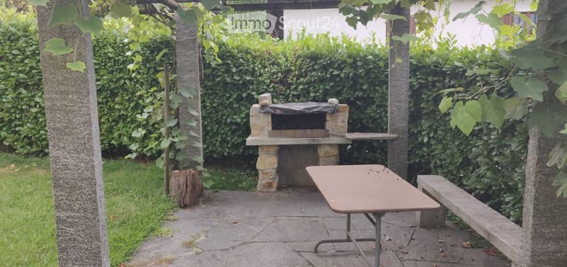 A small outdoor patio area with a stone barbecue, a wooden table, and a bench, surrounded by greenery and a hedge.