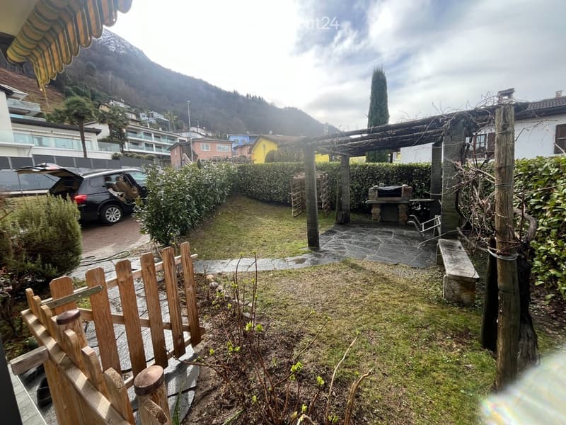 Garden with wooden fence, stone patio, barbecue, and surrounding greenery, with a car parked nearby and a mountain view in the background.