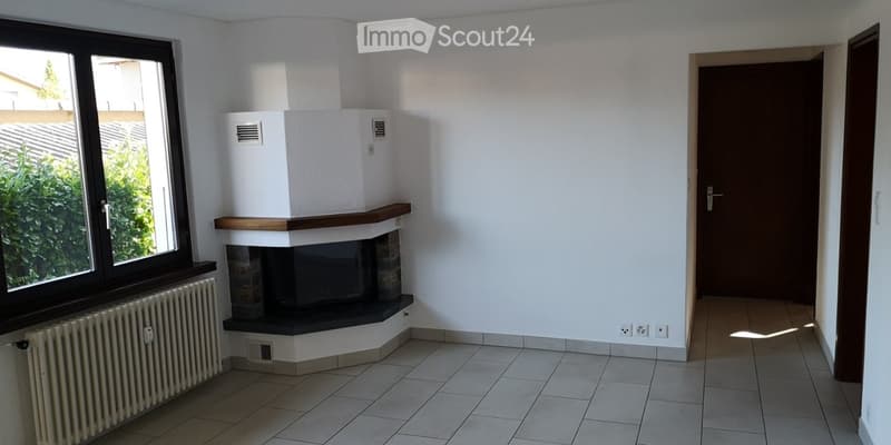 Empty room with tiled floor, white walls, fireplace, radiator, and large window with view of greenery.