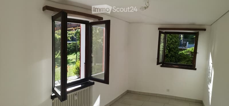 Empty room with white walls, tiled floor, and two windows with wooden frames. One window is open, revealing a view of a garden and trees outside.