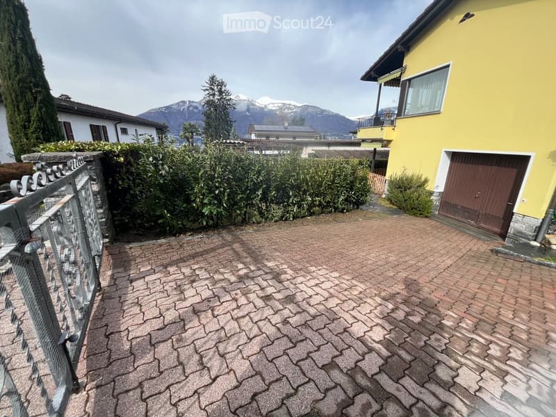 Brick-paved outdoor area with a metal fence on the left, a yellow house with a garage on the right, and a view of mountains and trees in the background.