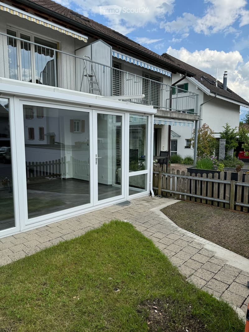 Garden area with a small lawn, paved pathways, a wooden fence, and a modern glass-enclosed patio connected to the house.